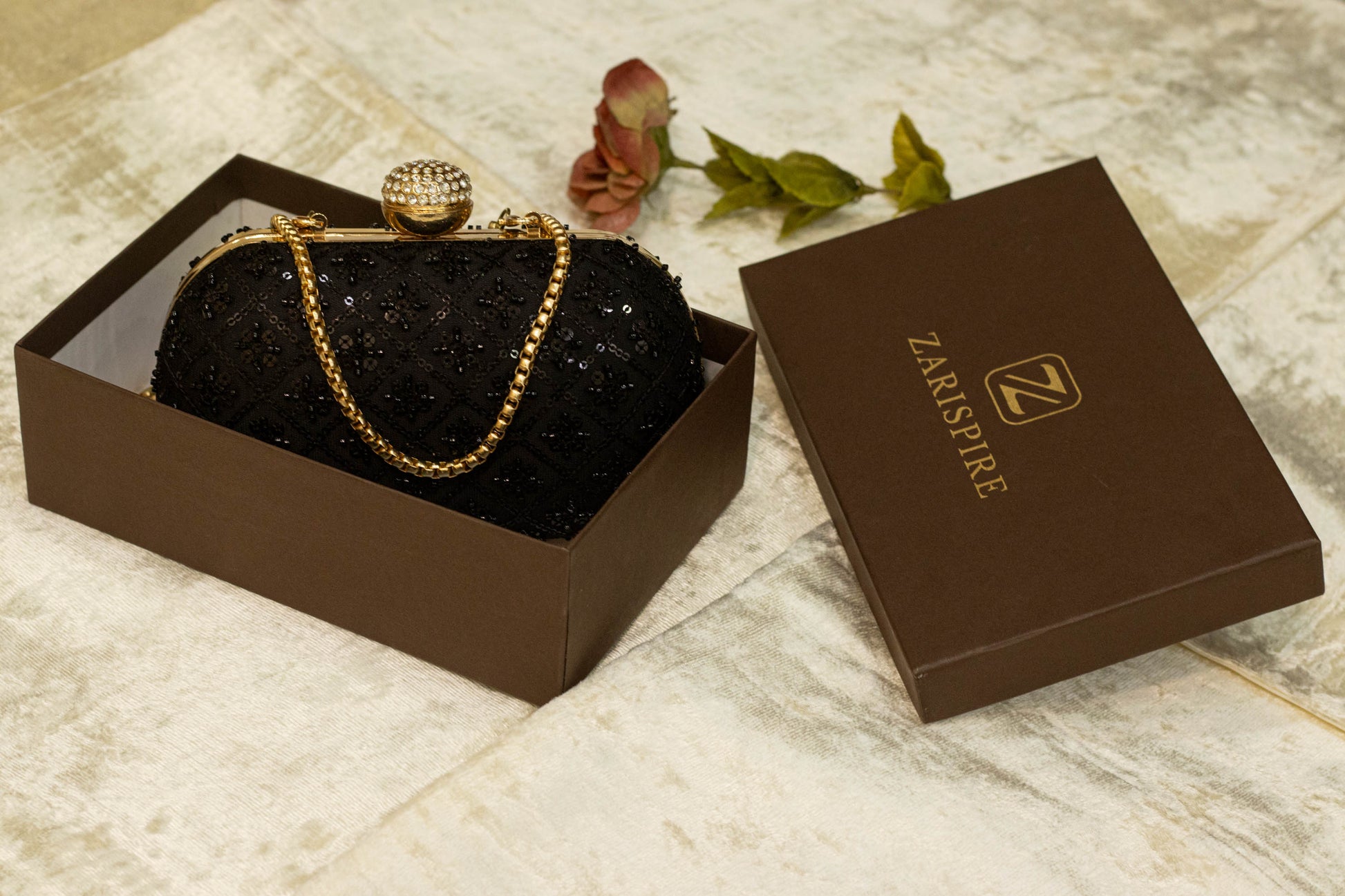 Black brocade Banarasi clutch bag with gold chain and spherical clasp in open brown box labeled Zari Sutra.