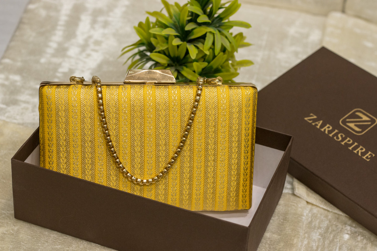 Yellow brocade Banarasi clutch bag with gold chain, traditional handmade design in a brown box.